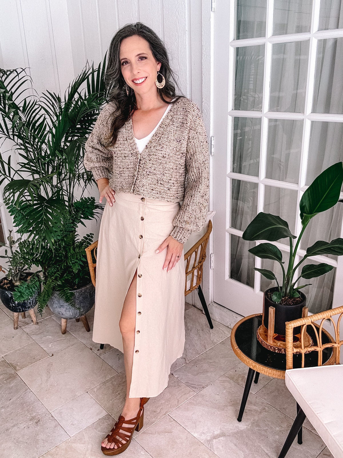 A woman in a beige skirt and knitted cardigan adorned with unique jewellery, posing by plants and a glass door, smiling at the camera.