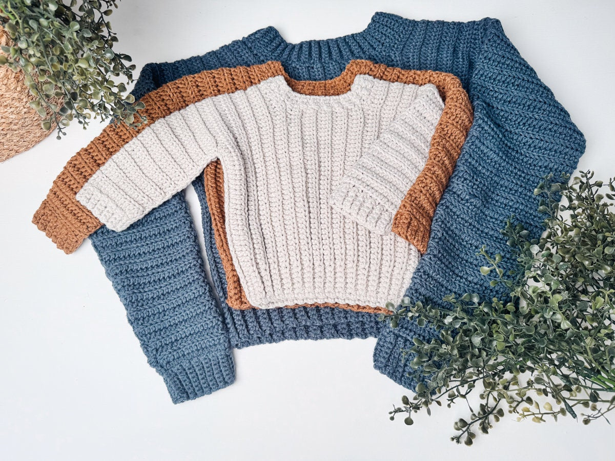 Three crocheted sweaters displayed flat, one in blue and the other in a cream, brown, and gray color-block pattern, surrounded by green foliage. Perfect as a handmade gift for any occasion.