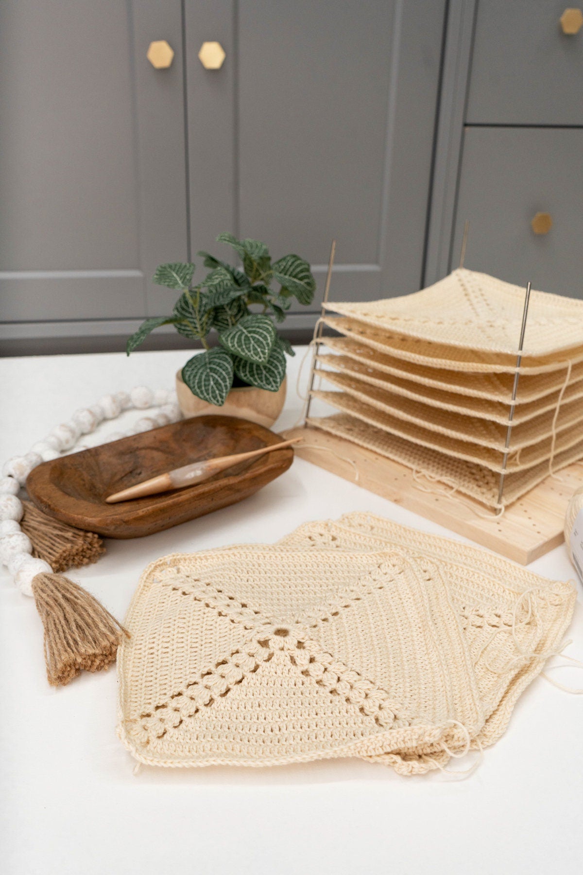 A display of crochet squares being stretched on blocking boards. Several completed squares, a wooden bowl with crochet tools, a string of beads, and a potted plant are on the table.