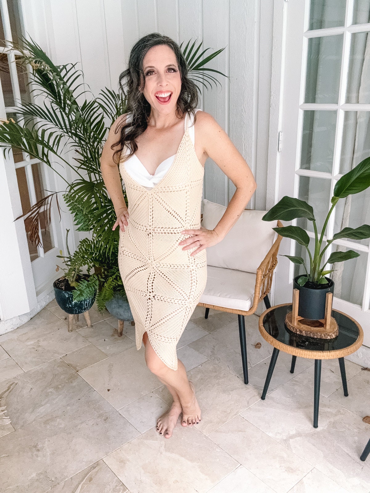 A woman stands barefoot indoors next to indoor plants, wearing a sleeveless, beige crochet dress and long, dark hair cascading over her shoulders. She poses with one hand on her hip, smiling at the camera.