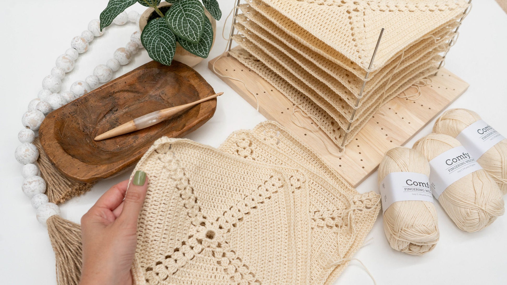A person holding a crocheted square next to multiple stacked crocheted pieces, three balls of yarn, a crochet hook, a potted plant, and decorative beads on a wooden tray—a perfect scene for crafting a handmade gift on a white surface.