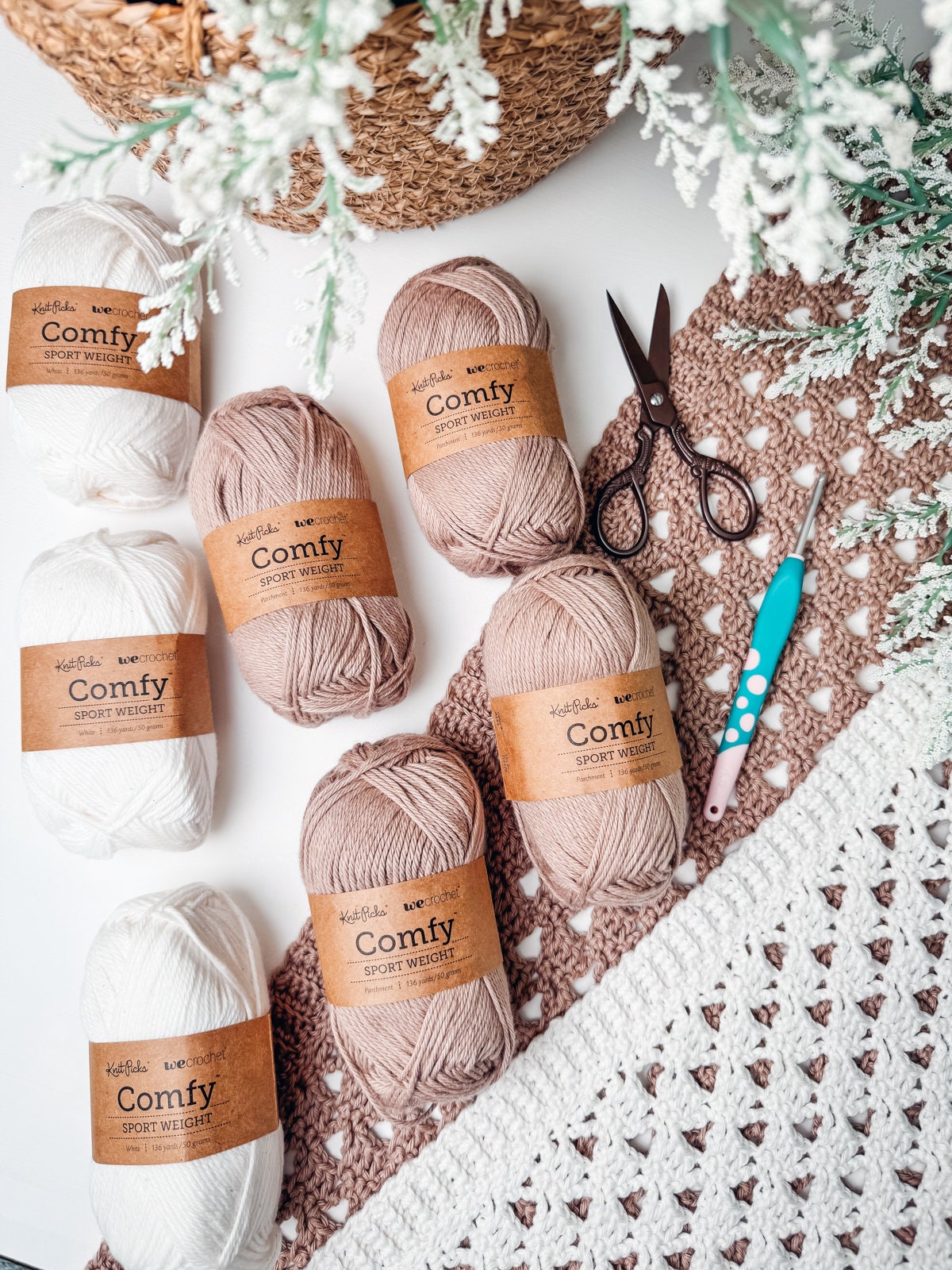 Yarn skeins labeled "Comfy Sport Weight" in cream, beige, and brown, a pair of scissors, and a crochet hook are placed on a partially crocheted garment, with flowers peeking out from a wicker basket surrounded by greenery.