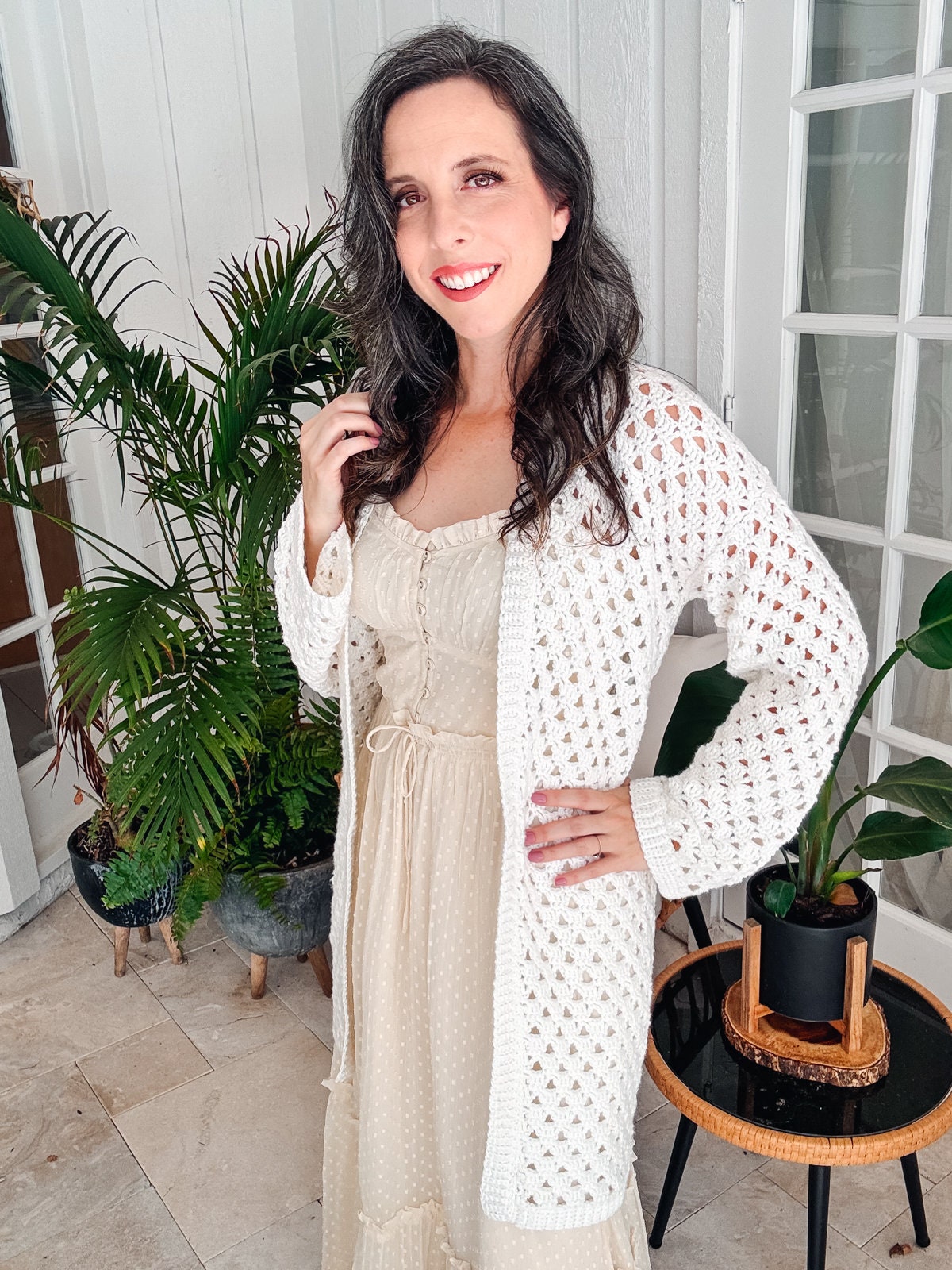 A woman with long, dark hair wearing a cream dress and a white crocheted cardigan stands indoors next to houseplants, accessorized with unique jewellery that perfectly complements her vintage clothes.