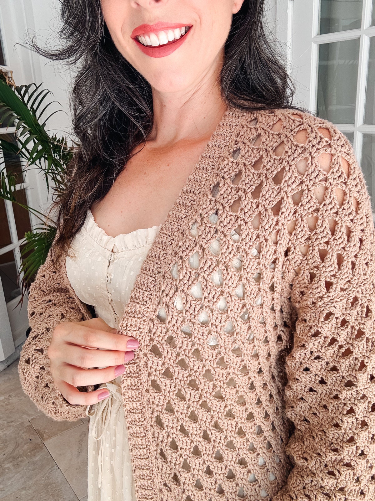 A person smiles while wearing a beige, crochet cardigan over a white dress, accessorized with unique jewelry. They are standing indoors beside a plant.