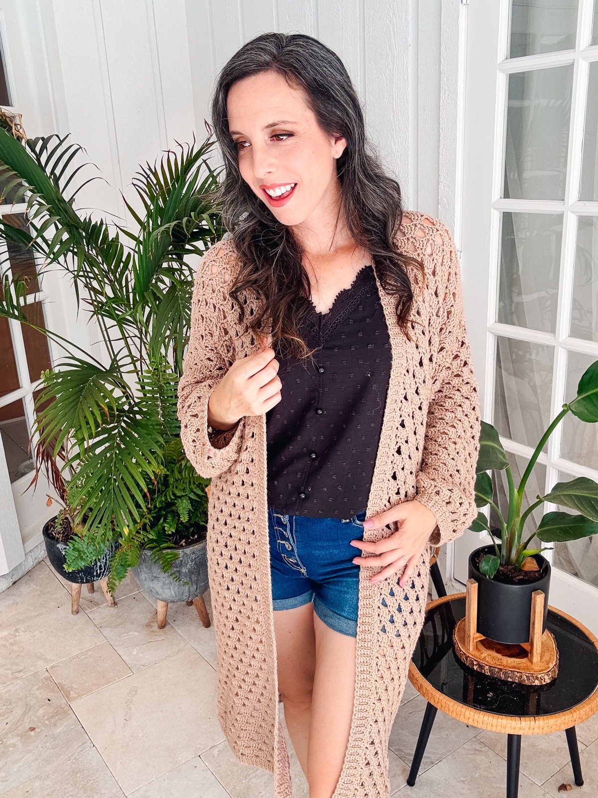 A woman with long, wavy hair stands indoors in front of plants. She is wearing a black top, blue shorts, and a beige open-crochet cardigan, she smiles and looks to her right.