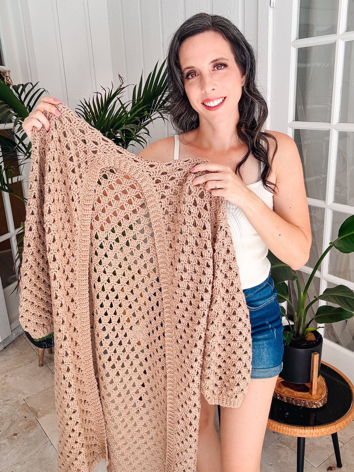 A woman in a white top and denim shorts holds up a beige crocheted cardigan in a well-lit room with plants in the background, showcasing her love for vintage clothes.