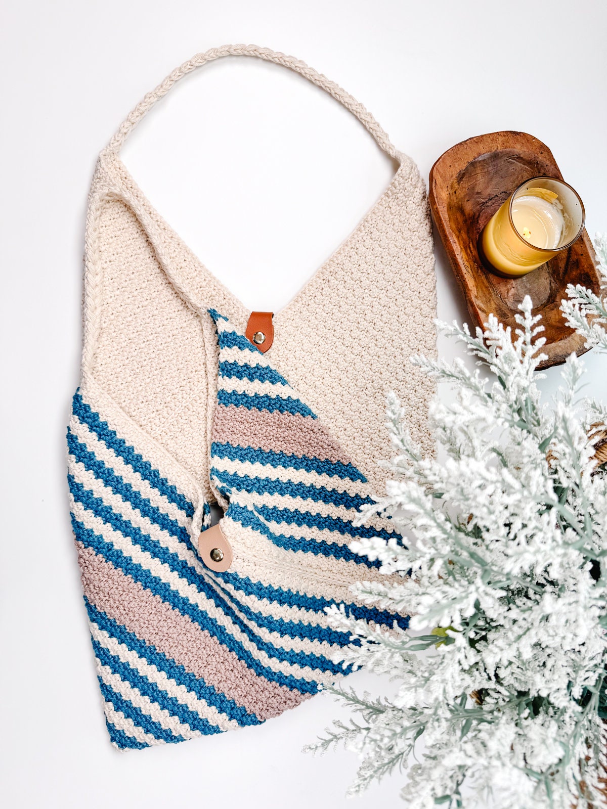 A crochet shoulder crossbody bag in beige and blue stripes is placed on a white surface. Next to it is a wooden tray holding a yellow candle and a frosted, evergreen branch arrangement, perfect as a handmade gift.