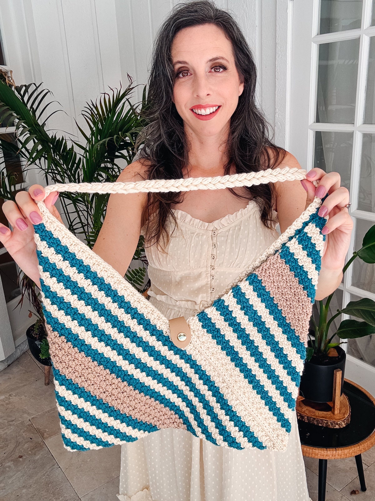 A woman smiles while holding a handmade crochet bag with diagonal stripes in shades of blue, white, and brown. She is standing indoors next to potted plants, her vintage clothes adding a charming touch to the scene.
