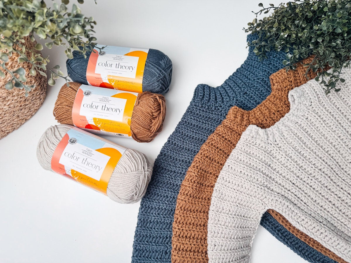 Three skeins of yarn in navy, brown, and beige are displayed next to crochet fabric swatches in matching colors, reminiscent of vintage clothes. Some greenery adds a touch of nature to the scene.