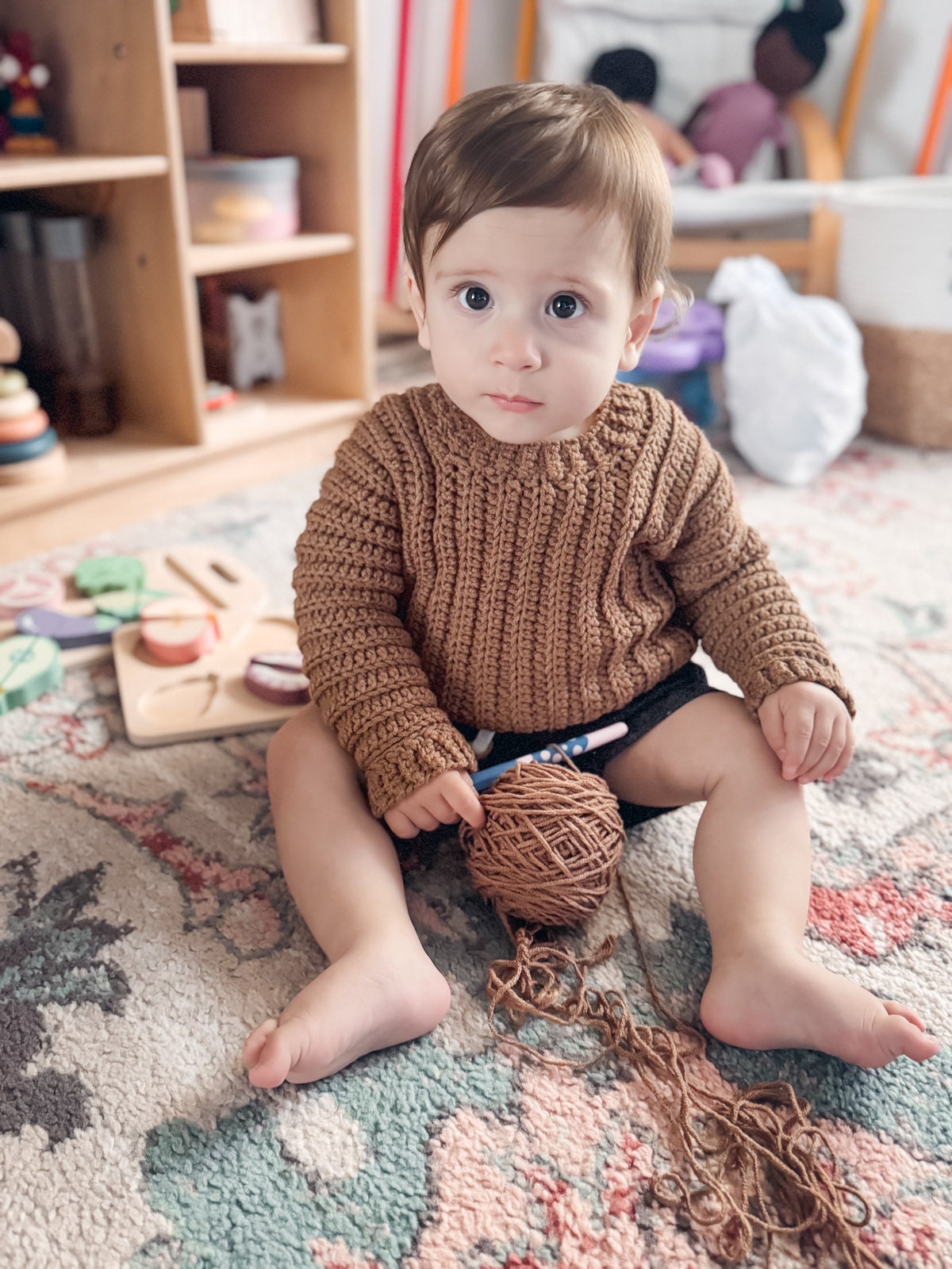 A baby sits on a patterned rug holding a ball of yarn, with toys and a bookshelf in the background. The baby wears a brown crocheted sweater that looks as cozy as vintage clothes from a handmade gift shop.