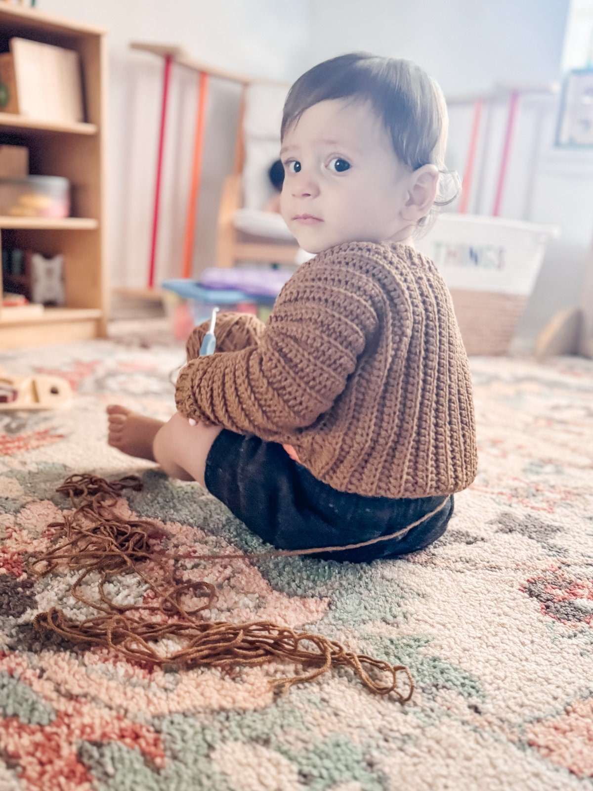 A baby wearing a brown crohcet sweater and black pants sits on a carpeted floor with scattered yarn, looking over their shoulder. Shelves adorned with unique toys and baskets in the background.
