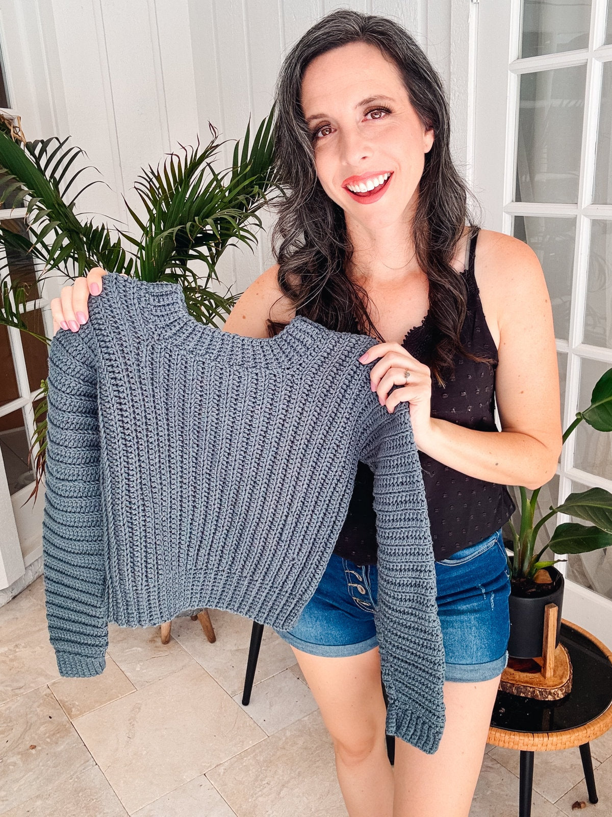 A smiling woman with long hair holds up a gray crochet sweater. She is wearing a black sleeveless top and denim shorts. There are potted plants behind her, and she seems to cherish the handmade gift, which adds a touch of charm to her look.