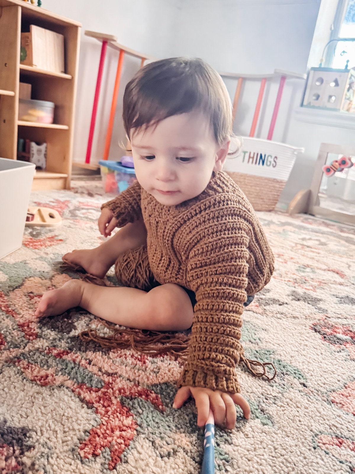 A young child in a brown crochet sweater sits on a patterned rug, reaching out. Shelves and toys are visible in the background, along with vintage clothes adorning the walls.