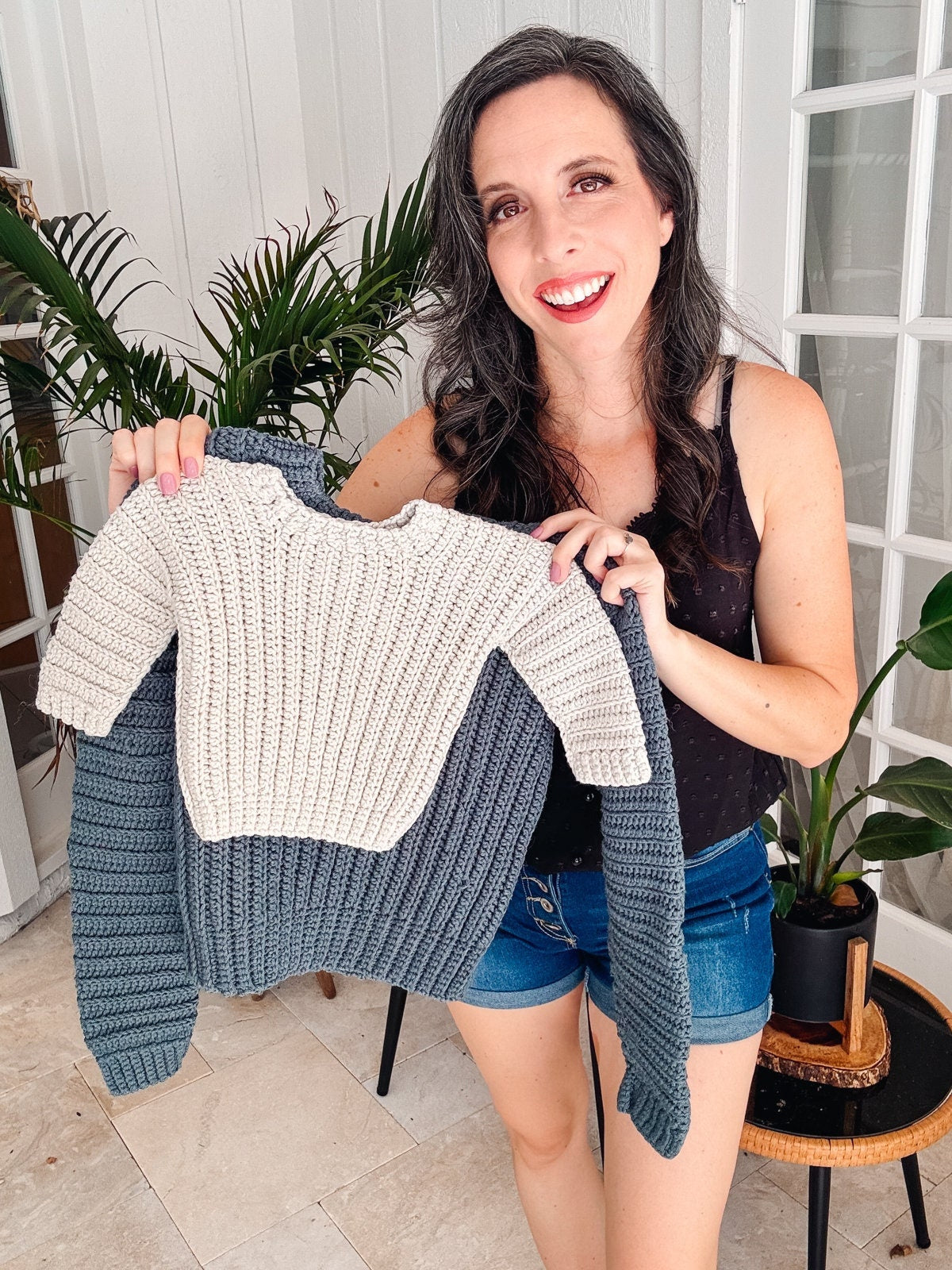 A woman in shorts and a sleeveless top holds up a color-blocked knit sweater, a perfect handmade gift. Indoor setting with plants in the background.