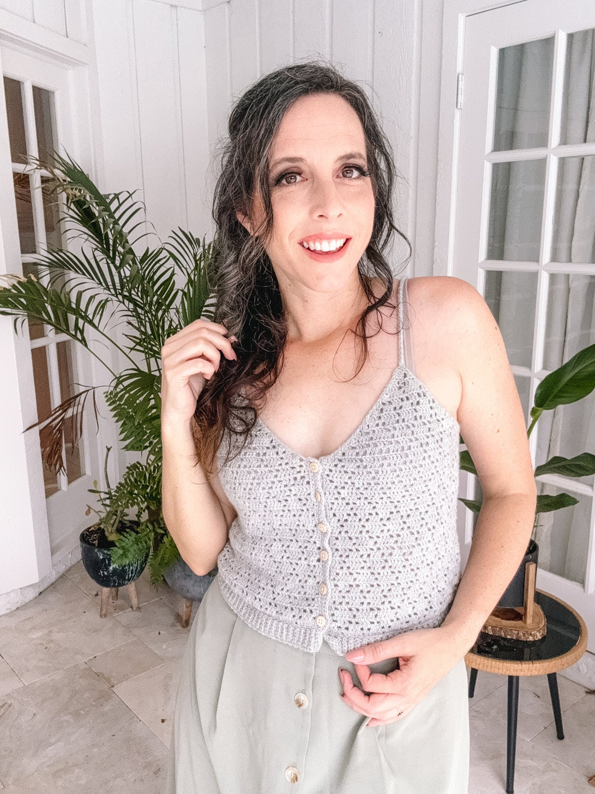 A woman with long, dark hair is smiling and standing indoors next to potted plants. She is wearing a light-colored, buttoned sleeveless top, a light green skirt, and unique jewellery that complements her vintage clothes perfectly.
