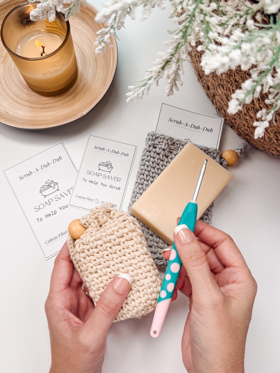 Hands holding a crochet soap saver with a crochet tool nearby, bar soap, a lit candle, and plant in the background. Cards with "Soap Saver" text visible suggest it as the perfect handmade gift.