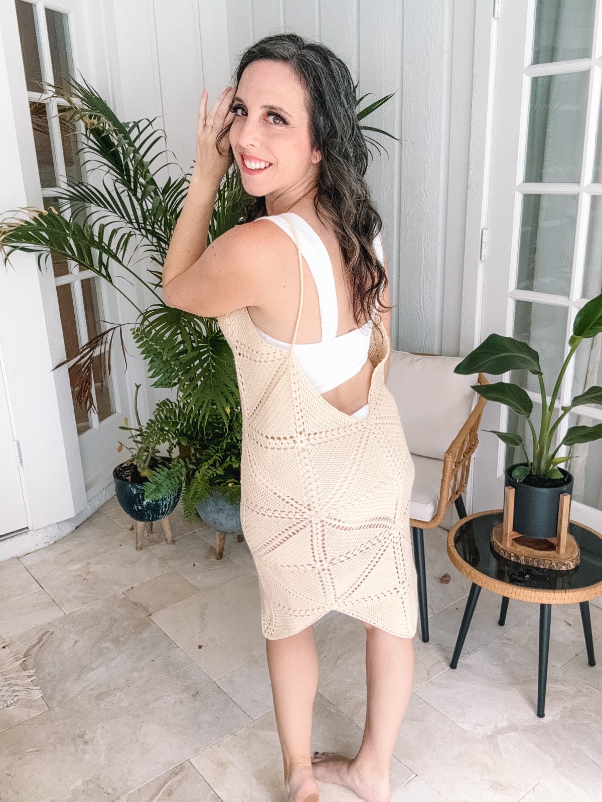 A woman stands indoors smiling and looking over her shoulder, wearing a light-colored crocheted dress over a white top. She is surrounded by potted plants and stands on tile flooring.