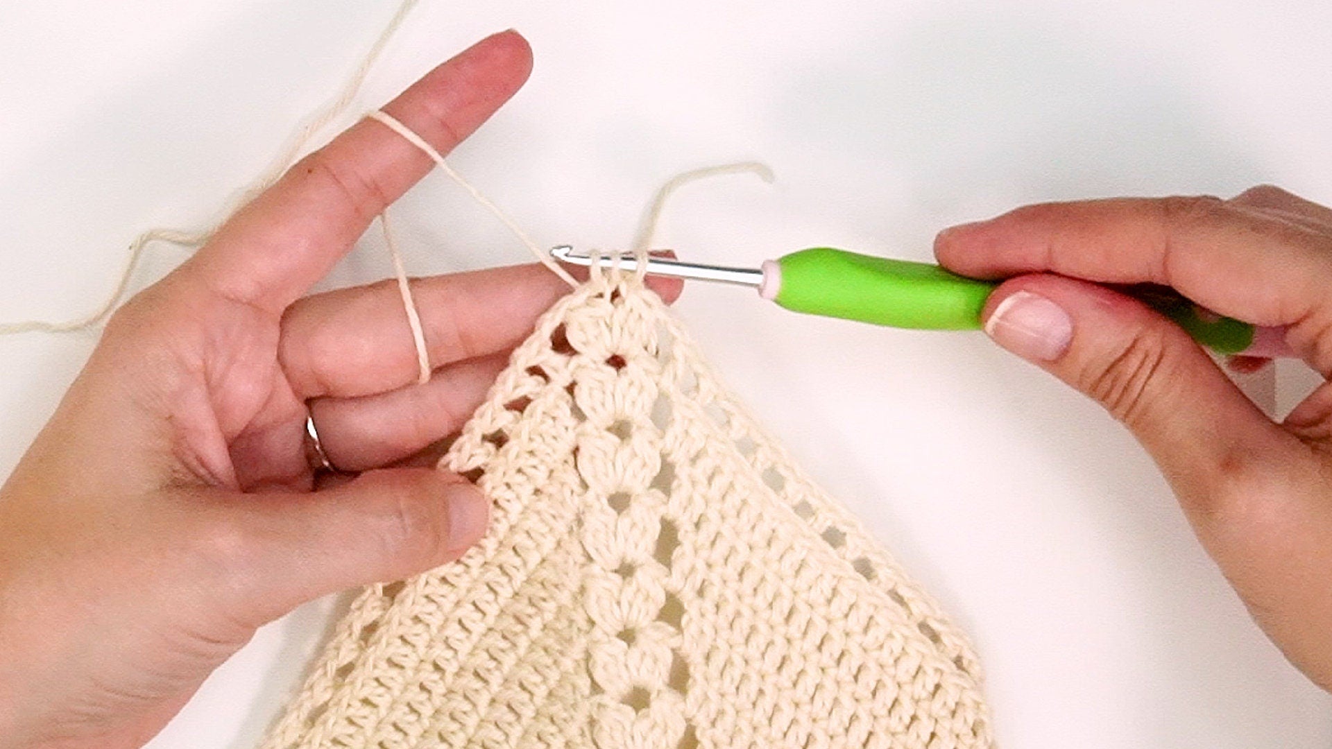 A person is crocheting a light-colored fabric using a green-handled crochet hook, perfect for creating a handmade gift. The fabric features a textured stitch pattern.