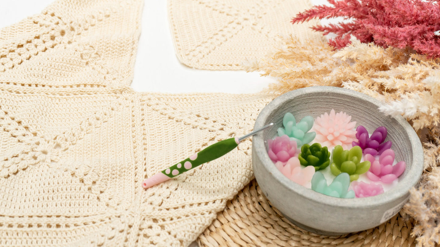 A crochet hook in a bowl with colorful crocheted succulent plants on a textured crochet fabric background, accented by pink and beige dried flowers.