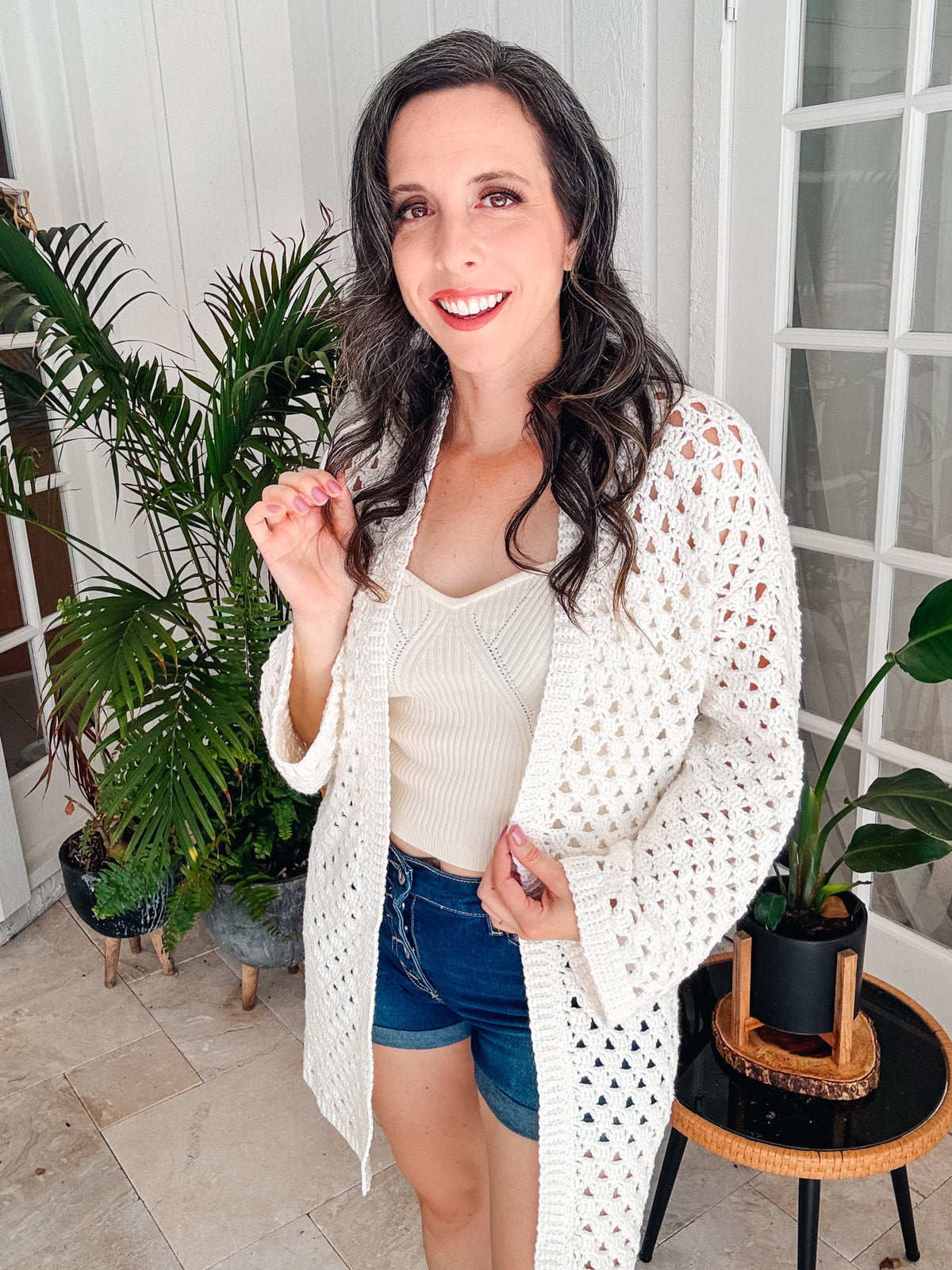 A woman with long dark hair wearing a white crochet cardigan, white top, and blue denim shorts stands indoors in front of a plant and a glass door, smiling at the camera.