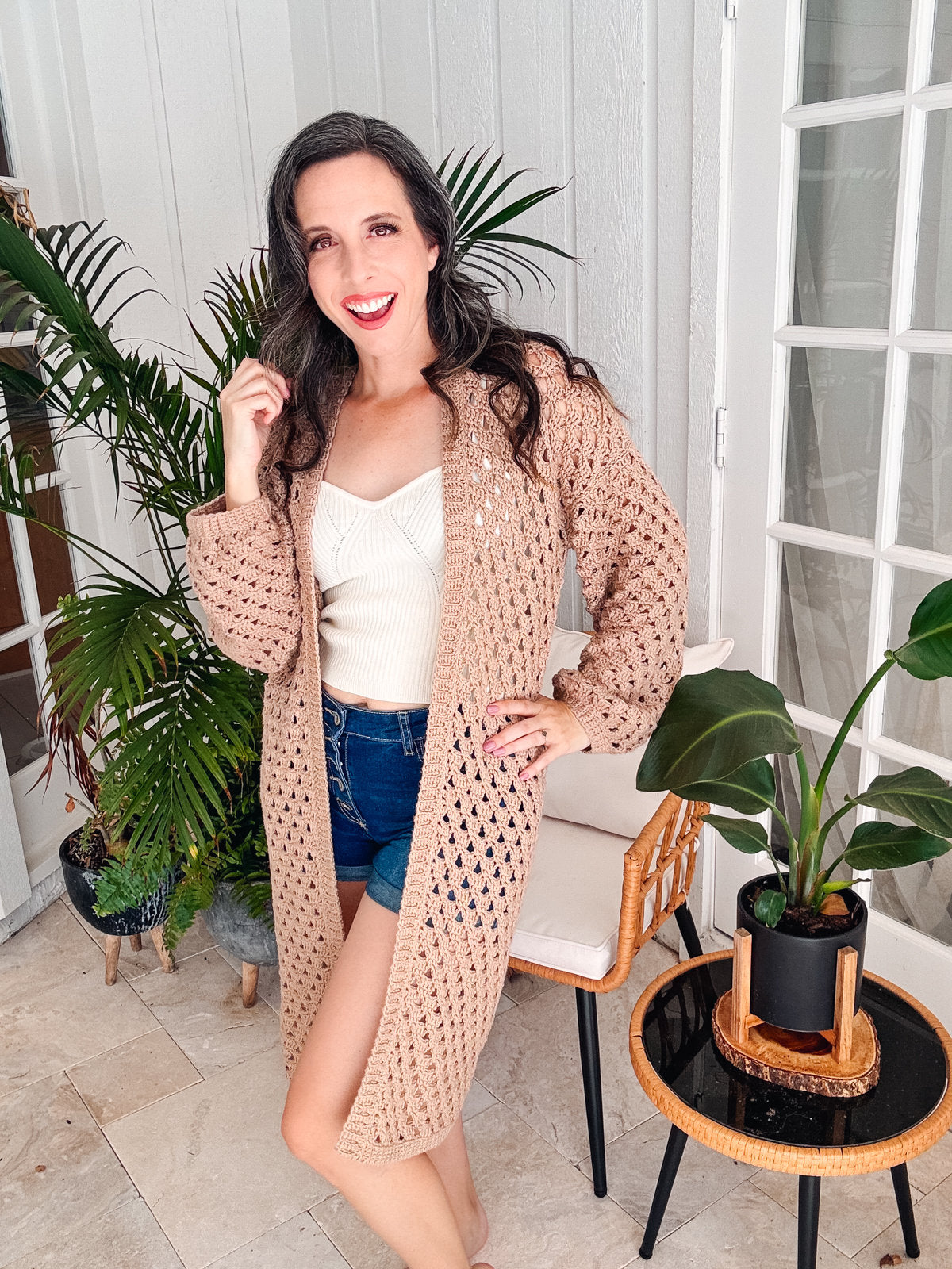 A woman stands indoors smiling, wearing a beige crochet cardigan over a white top and denim shorts. She is posing near a small plant and a round side table, with large green plants in the background.
