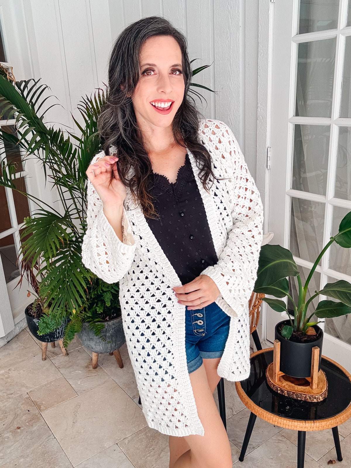 A woman wearing a white crocheted cardigan over a black top and denim shorts. She is posed smiling, with one hand on her hip and the other slightly raised. There are potted plants in the background, adding a touch of greenery to the scene.