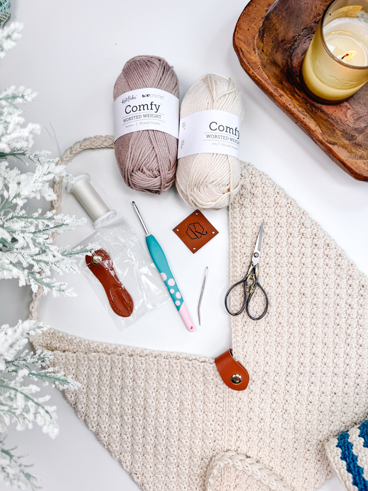 A flat lay image displays crochet tools including yarn balls, crochet hooks, a pair of scissors, a button, and a label. Also visible are a small dish with a candle and a portion of a crocheted piece, perfect for crafting unique handmade gifts.