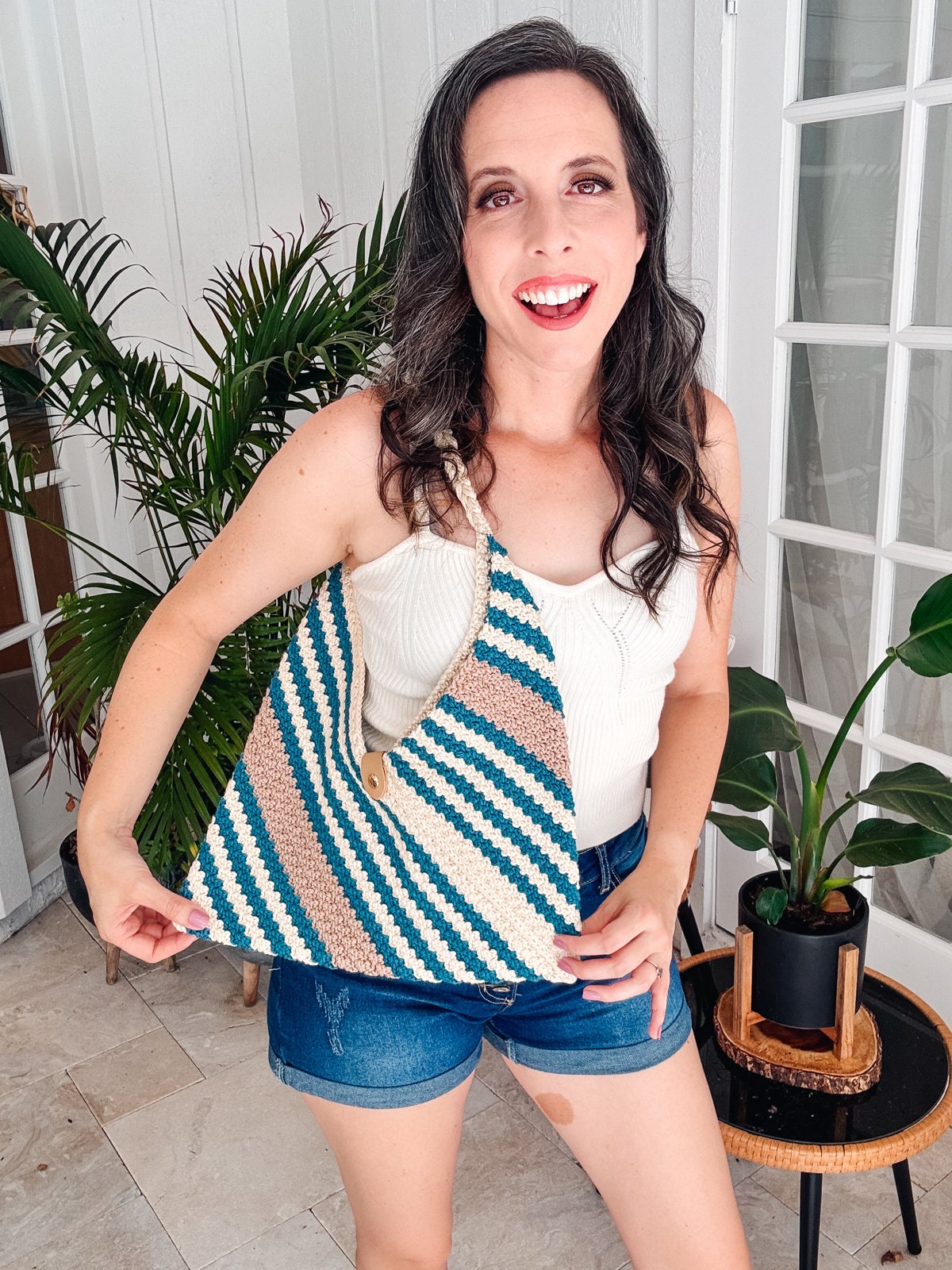A woman in a white top and denim shorts smiles while holding a striped crocheted bag, which she's considering as a handmade gift. Indoor setting with potted plants in the background.