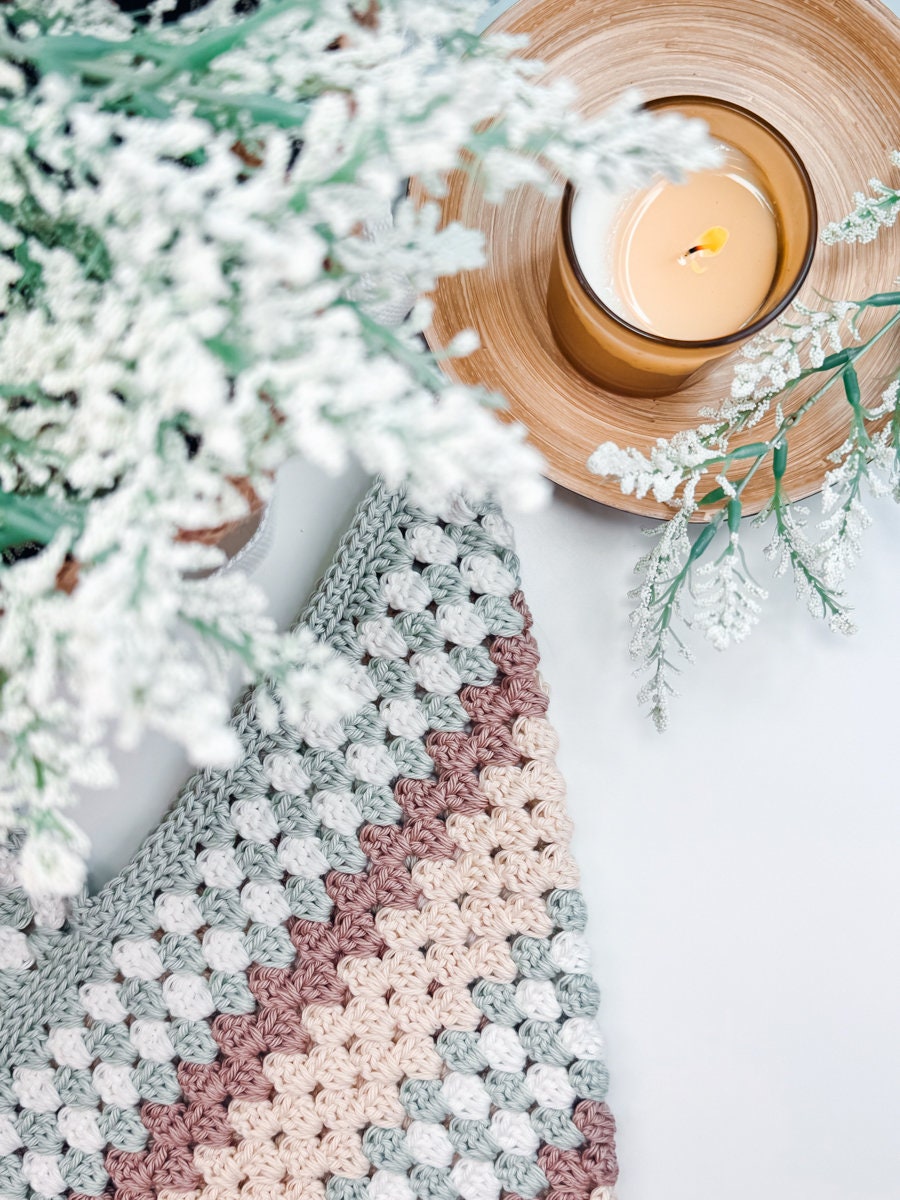 A lit candle in a wooden holder next to a plant with white flowers and a multicolored crocheted cloth, making for an ideal scene for showcasing a crocheted bag.
