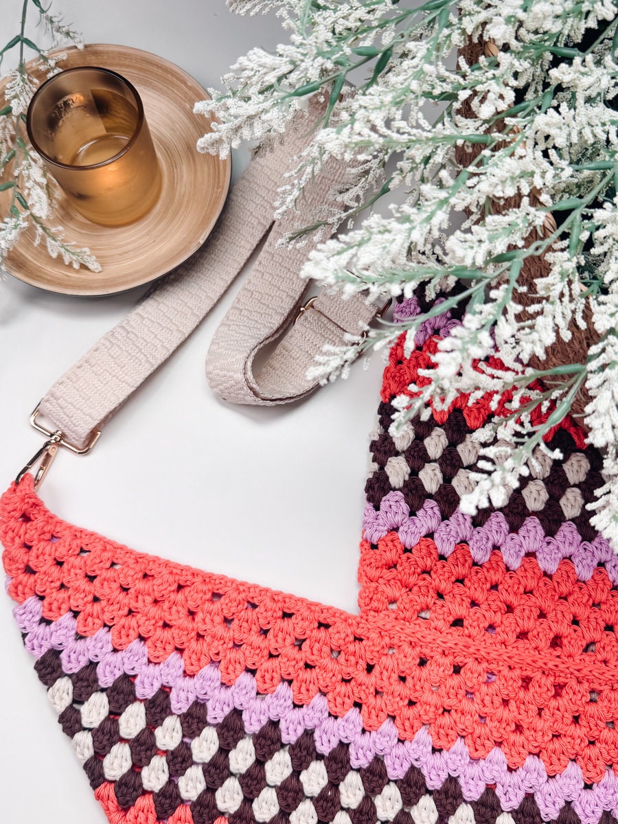 A crocheted bag with pink, purple, and brown stripes, like the Taylor Swift Dress, sits next to a plant with frosted leaves and a lit candle in a glass holder on a circular wooden tray, making for an ideal handmade gift.