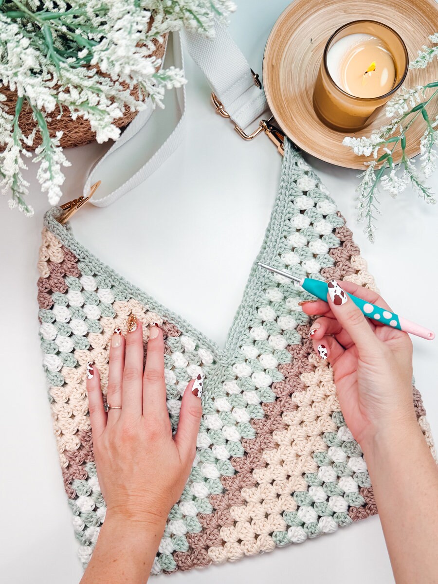 Hands crocheting a triangular pattern bag with pastel-colored yarn. The scene includes a lit candle and green decorative plants. The person is using a blue-handled crochet hook, crafting what could be an ideal handmade gift.