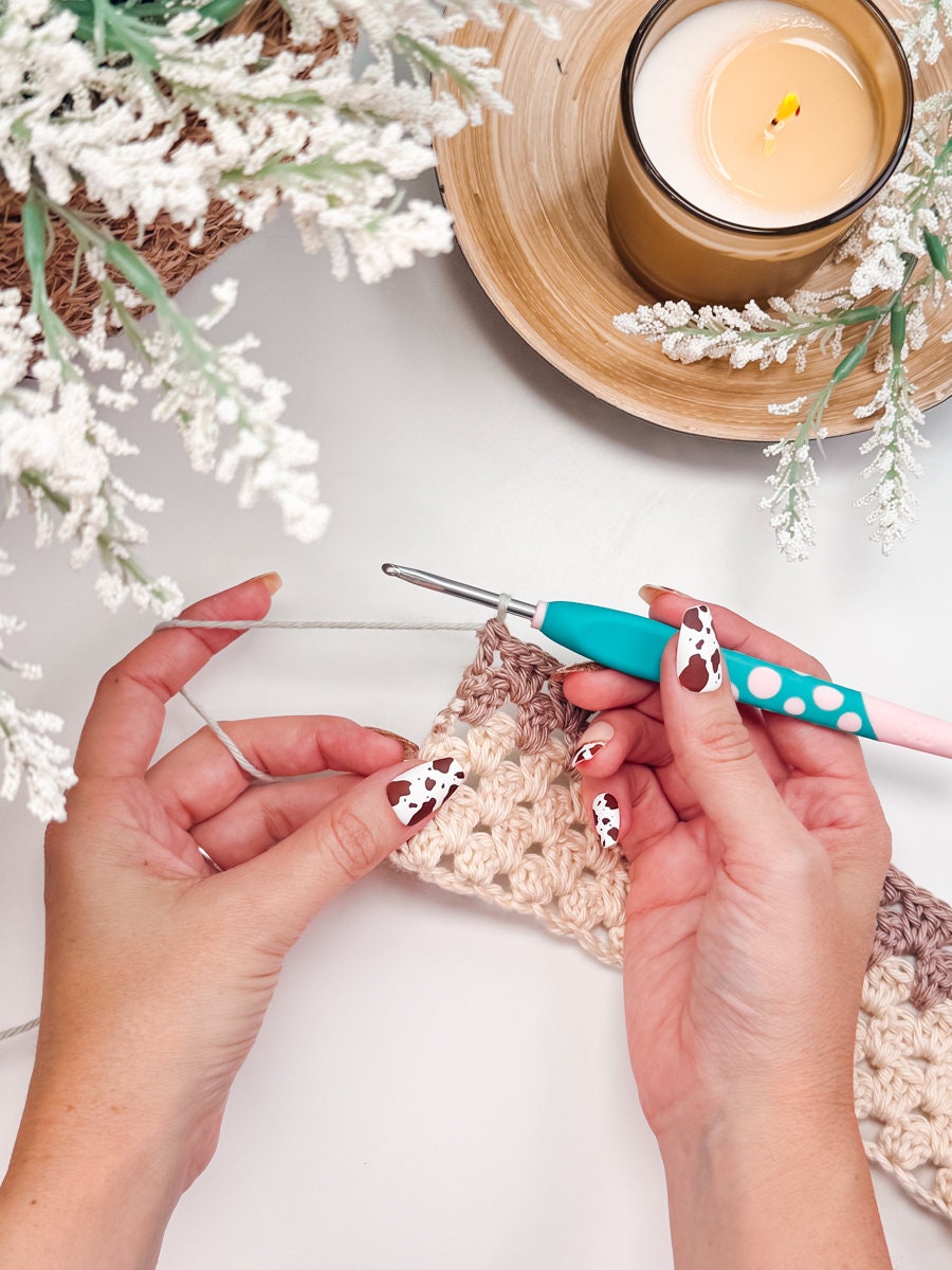 A person crochets with a teal and pink hook while holding a piece of yarn, creating a handmade gift. Nearby, a lit candle casts a warm glow on the white floral decor.