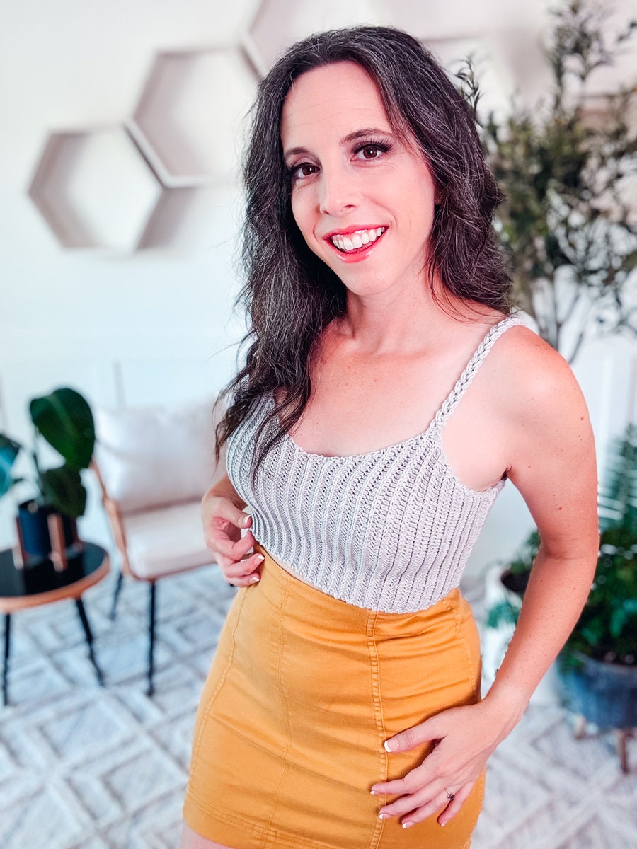 A woman with long dark hair wearing a light gray crochet tank top and a mustard yellow skirt stands indoors, showcasing unique jewelry, with hexagonal shelves and plants in the background.