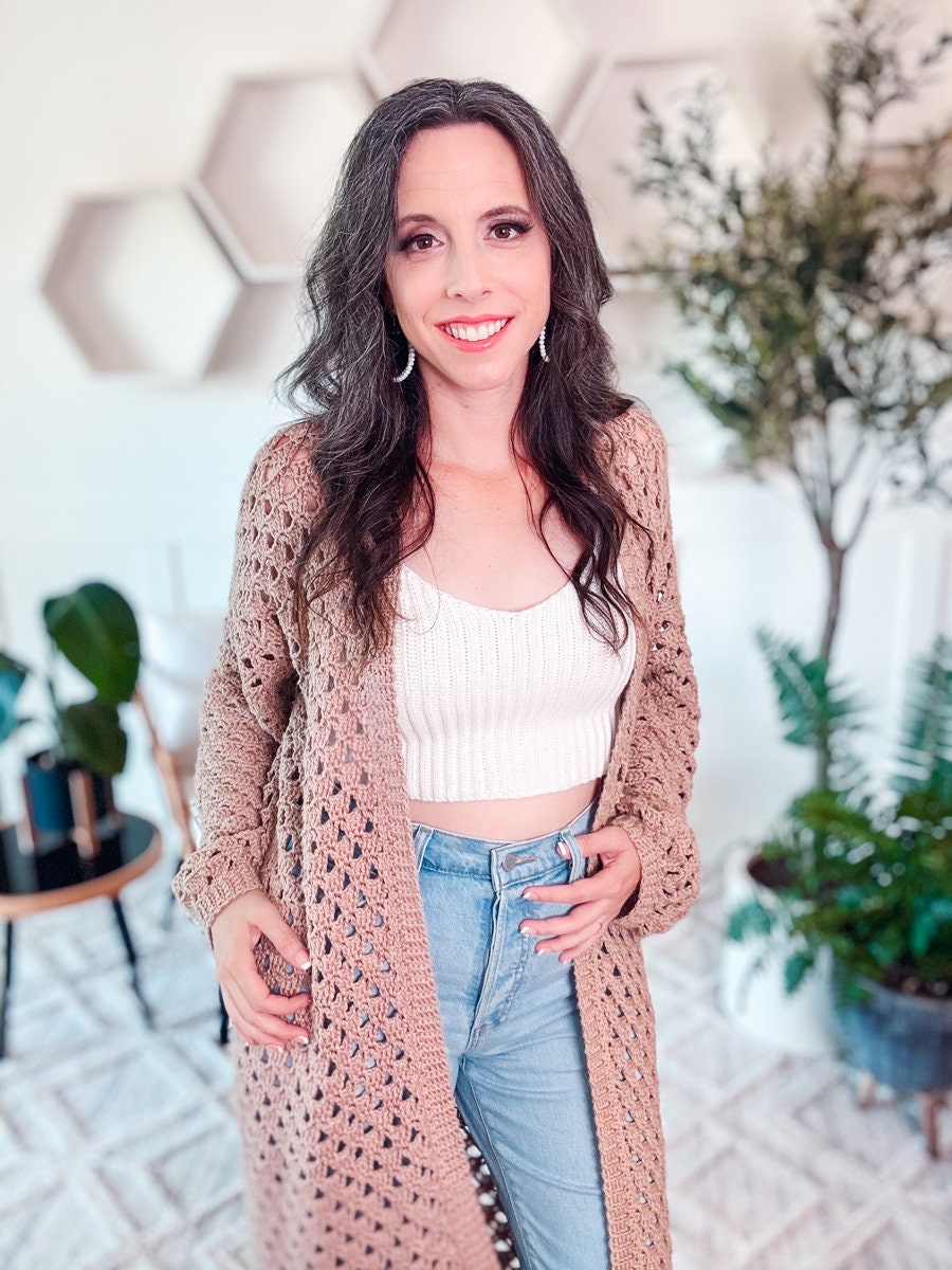 A woman with long dark hair wearing a white crochet crop top, blue jeans, and a beige crochet cardigan stands indoors in a room adorned with plants and geometric wall decor.