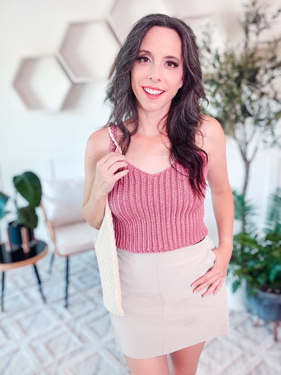 A woman with long, dark hair is smiling, wearing a sleeveless pink crochet top and a light-colored skirt, holding a beige bag. She is standing in a room with hexagonal wall decor and indoor plants.
