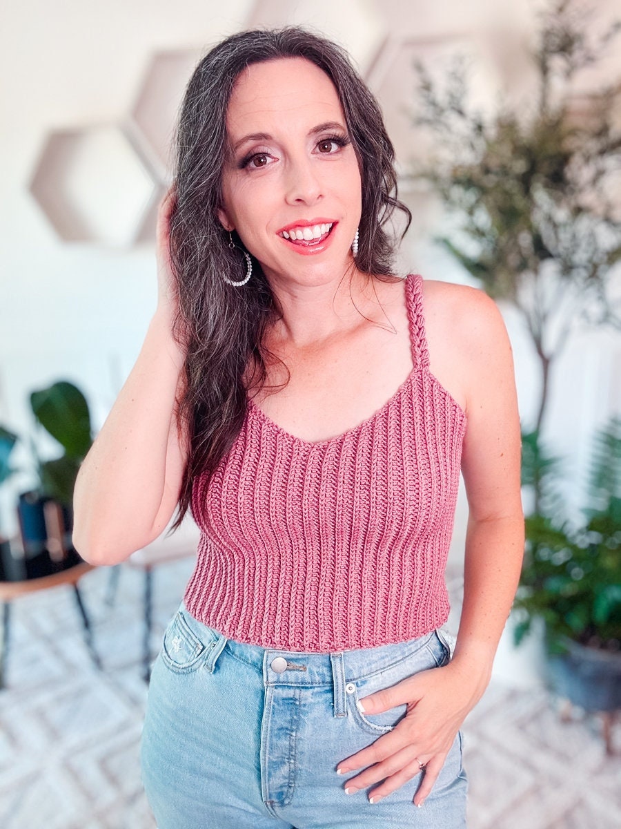 A person with long dark hair wearing a red knitted tank top and blue jeans, standing indoors with one hand touching their hair, smiling, and showcasing their unique jewelry.