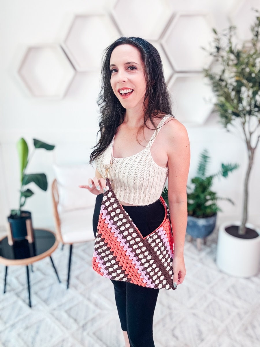 A woman stands indoors, showcasing a granny stitch crochet shoulder bag with a colorful pattern. She is wearing a sleeveless top and black pants. The background features hexagonal wall decor and potted plants.
