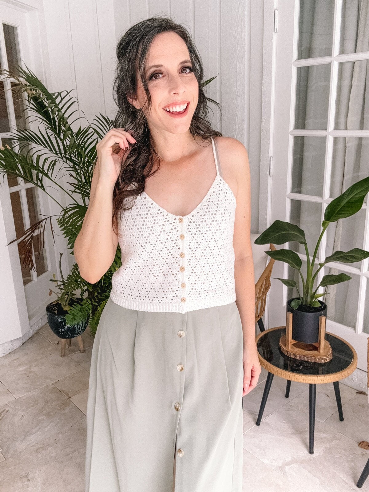 A woman stands indoors, smiling at the camera. She is wearing a white knitted sleeveless top and a light green buttoned skirt. There are plants and a small table in the background.
