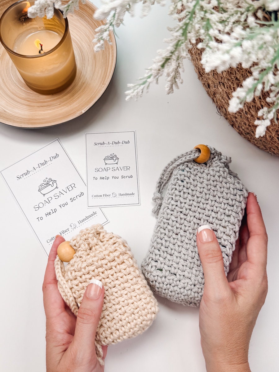 Hands holding two crocheted soap savers with product tags that read "Soap Saver to help you scrub." A lit candle and white flowers are in the background, making these handmade gifts perfect for anyone who loves a touch of cozy elegance.