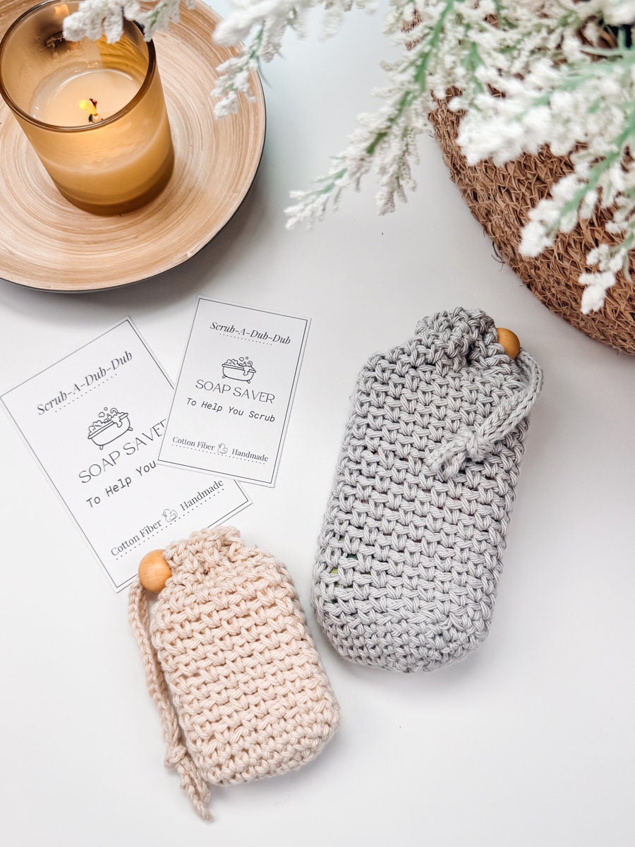 Two crocheted soap savers, one beige and one gray, placed next to a scented candle and instruction cards on a white surface make for a perfect handmade gift.