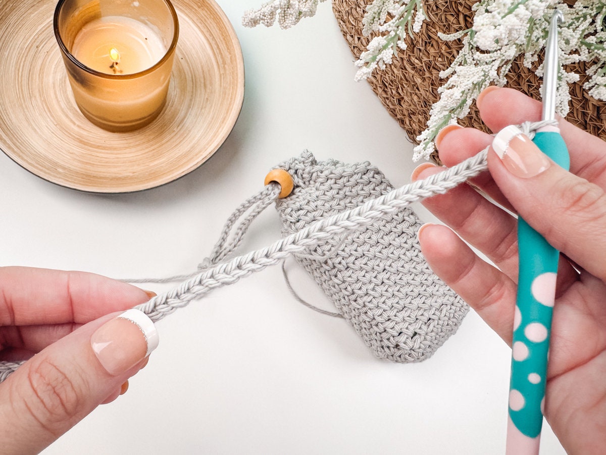 Hands holding a crochet project with a blue polka dot hook next to a lit candle on a wooden coaster, alongside a knitted pouch in the background, evokes the charm of crafting a heartfelt handmade gift.