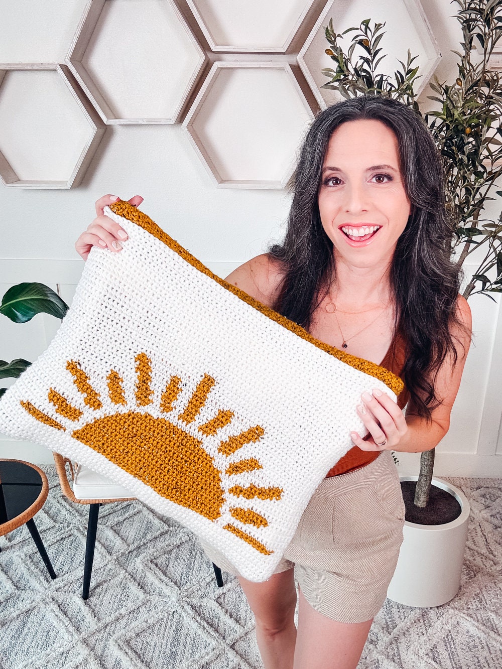 A person holding a crocheted blanket with a sun design. The background includes honeycomb-shaped wall decor, an indoor plant, and a stool adorned with unique jewellery, adding charm to the cozy setting.