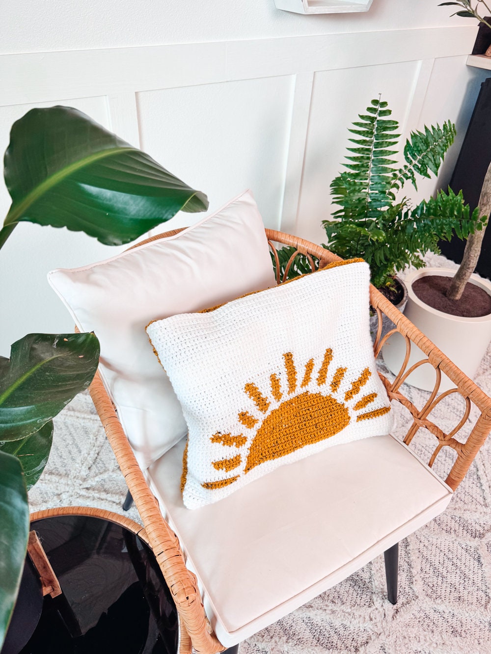 A rattan chair with a white cushion and a knitted pillow featuring a yellow sun design sits surrounded by green potted plants, making the perfect spot to unwind while browsing unique jewellery or vintage clothes online.