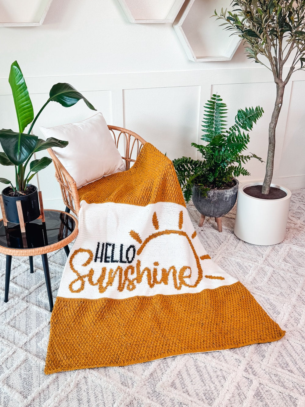 A wicker chair with a mustard yellow and white blanket draped over it, featuring the words "Hello Sunshine" and a sun illustration. The surrounding area includes potted plants.
