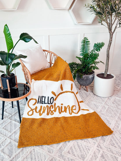 Mustard and white crochet blanket with a sun and lettering draped over a wicker chair among potted plants