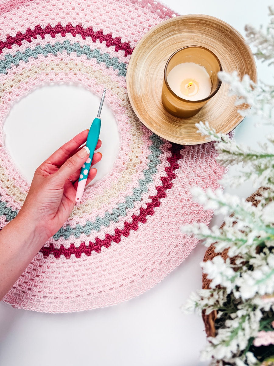A hand holding a crochet hook over a circular crochet project with striped colors. A lit candle in a wooden bowl and a frosted pine branch are also visible, evoking the charm of handmade gifts.