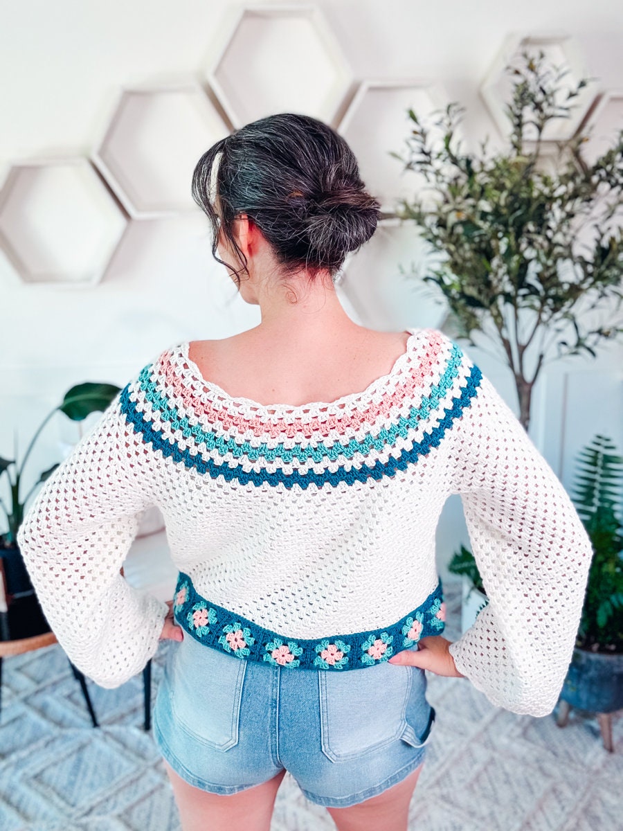 Person facing away, wearing a white crochet sweater with colorful patterns and denim shorts. Background features hexagonal wall decor and plants, evoking the charm of vintage clothes.