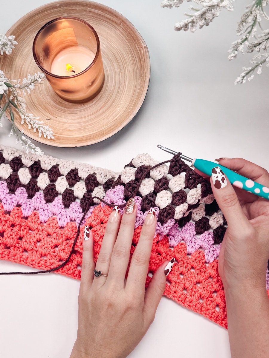 A close-up of hands crocheting a colorful striped fabric with a crochet hook, next to a lit candle in a wooden tray. The person’s nails, decorated with a cow print design, hint at the creation of a handmade gift.