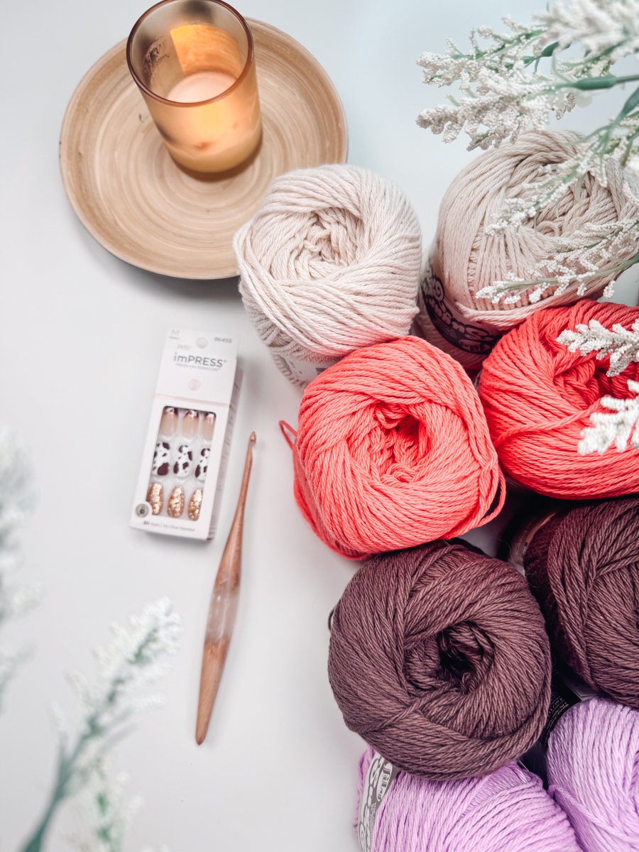 A collection of yarn balls in various colors, a wooden crochet hook, unique jewellery, a pack of artificial nails, and a lit candle on a wooden coaster, surrounded by green foliage.