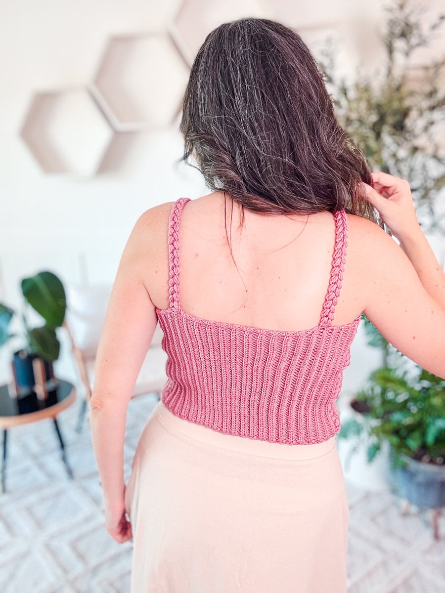A person with long hair facing away, wearing a pink crocheted tank top and beige skirt. The background shows a modern indoor setting with plants and hexagonal wall decorations.